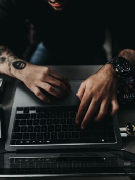 Close-up of tattooed hands with a wristwatch typing on a laptop keyboard, symbolizing modern tech lifestyle.