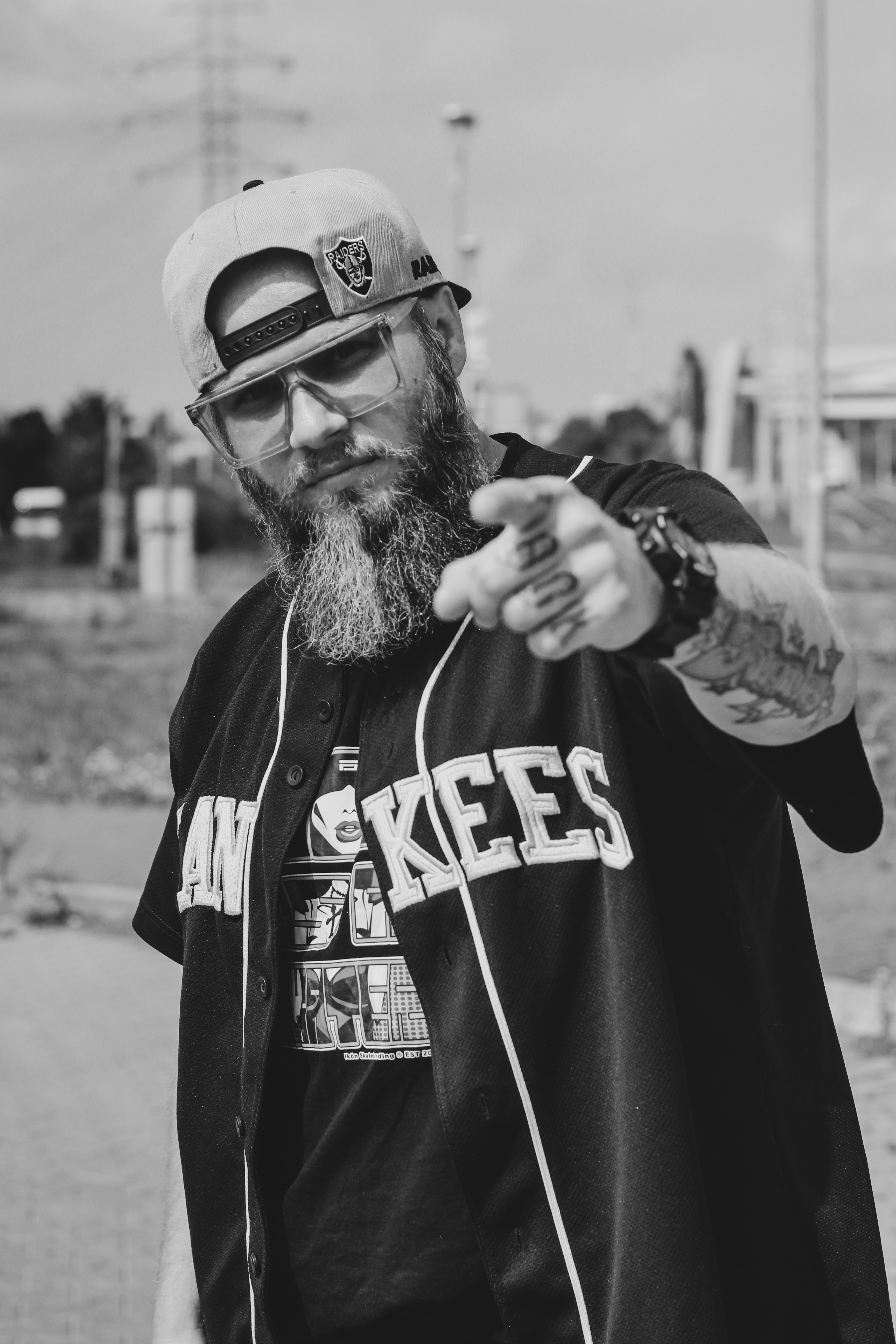 Black and white photo of a bearded man in a street style outfit pointing ahead.