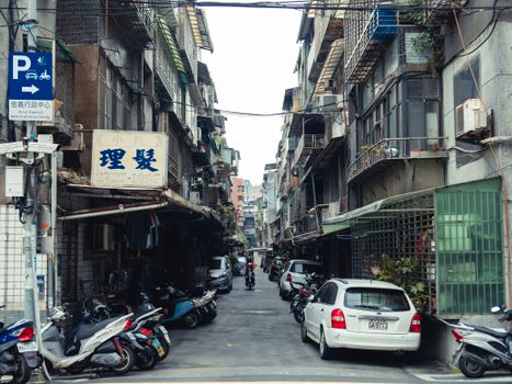 Narrow urban alley with parked cars and scooters in an Asian city, showing lively street life.