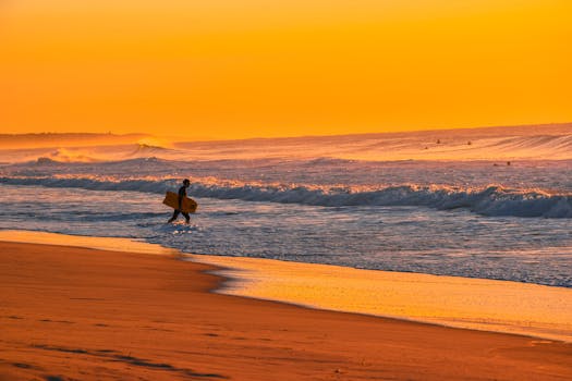 Surfer walking along sandy beach during a vibrant orange sunset, creating a tranquil seascape.
