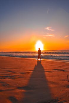 A serene silhouette of a woman walking on a sandy beach during a vibrant sunset, creating a peaceful scene.