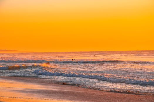 Stunning golden sunset over the ocean with gentle waves and surfers on the horizon.