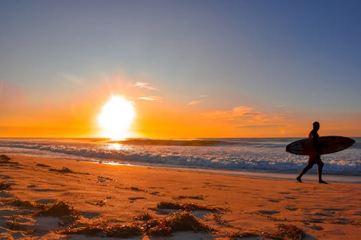 Silhouetted surfer walking on the beach during a vibrant sunset, creating a peaceful seascape scene.