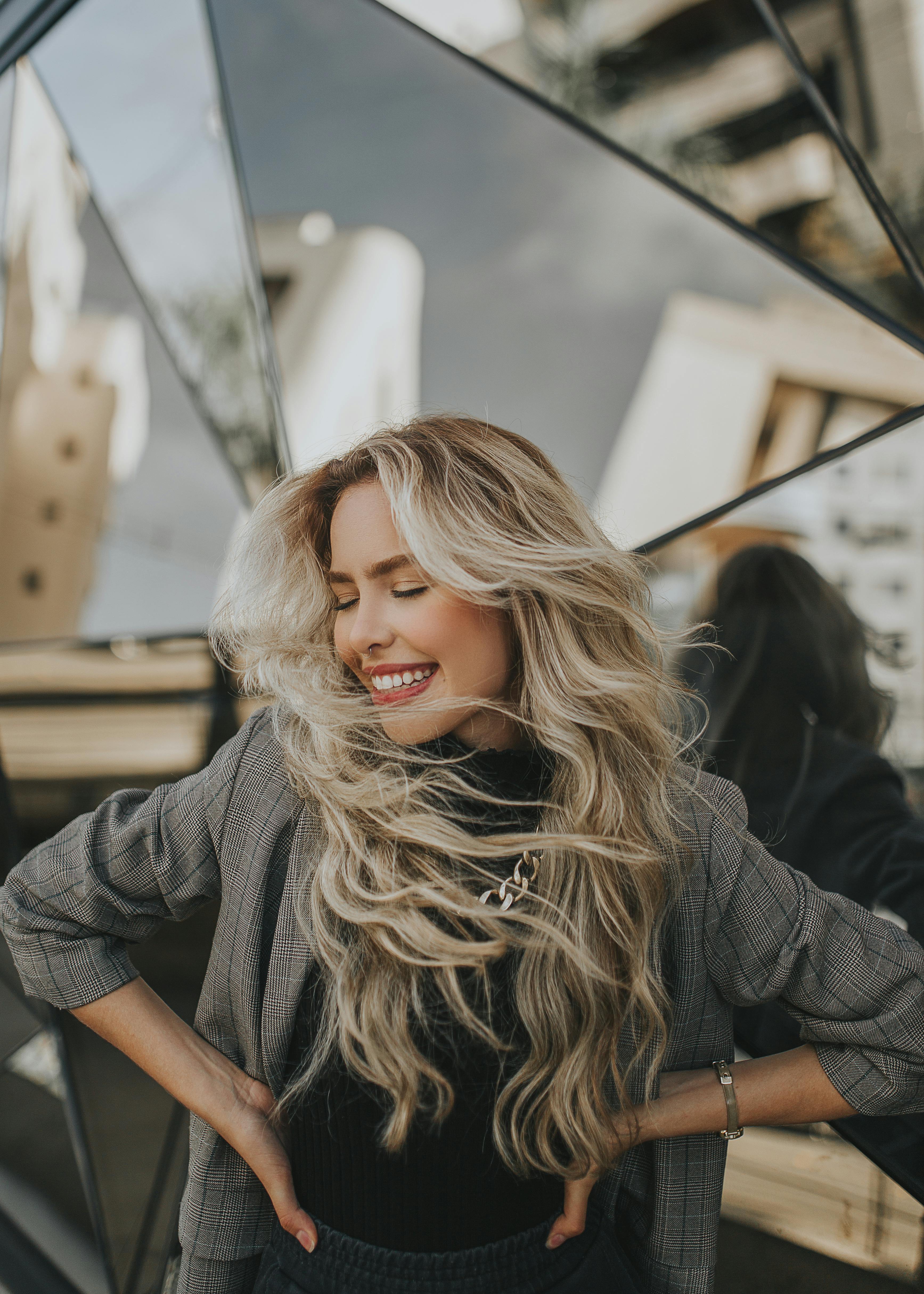 Blonde woman in fashionable checked jacket posing with a smile and windswept hair in an urban setting.