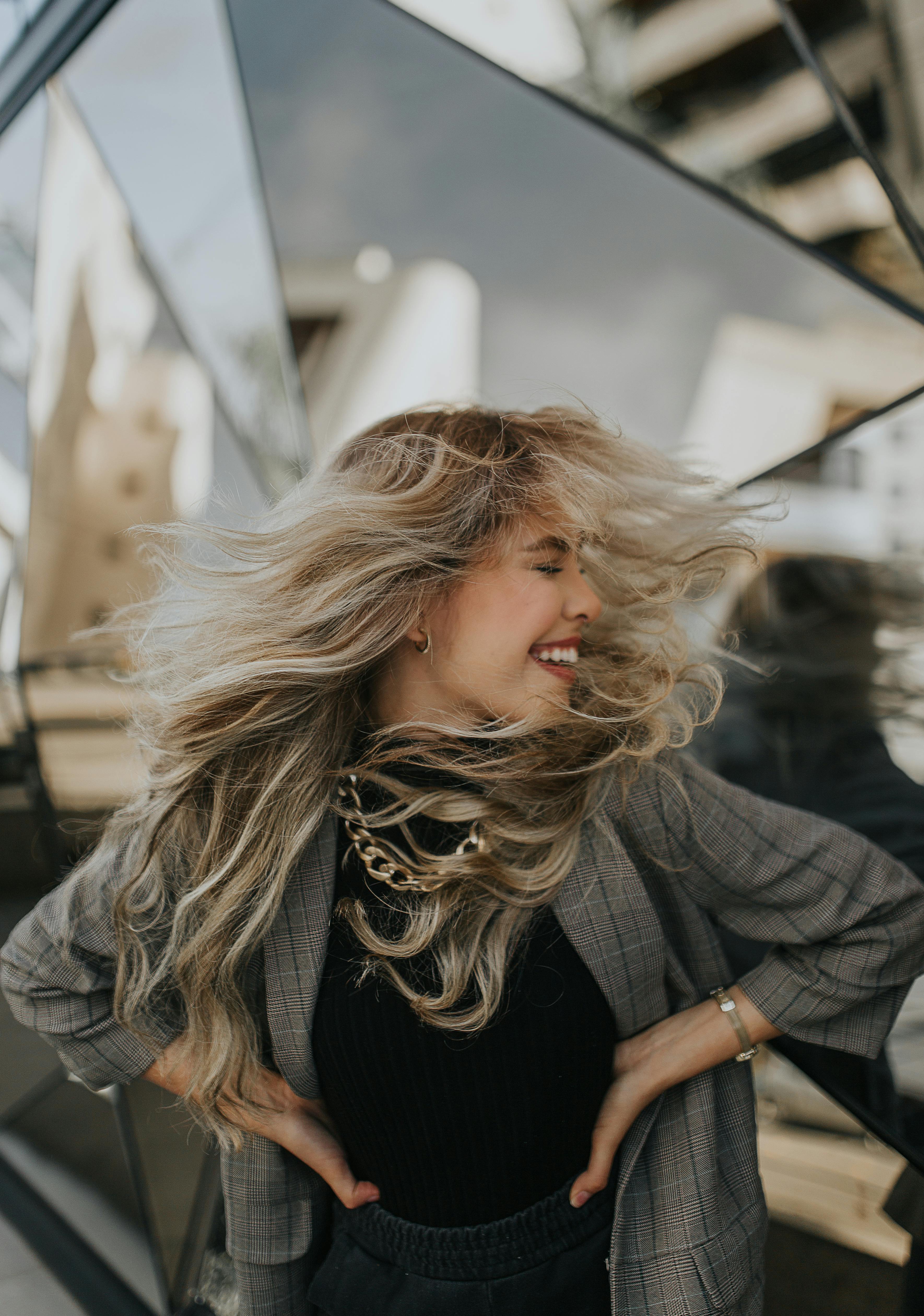 Blonde woman in fashionable attire joyfully tosses her hair against a modern urban backdrop.