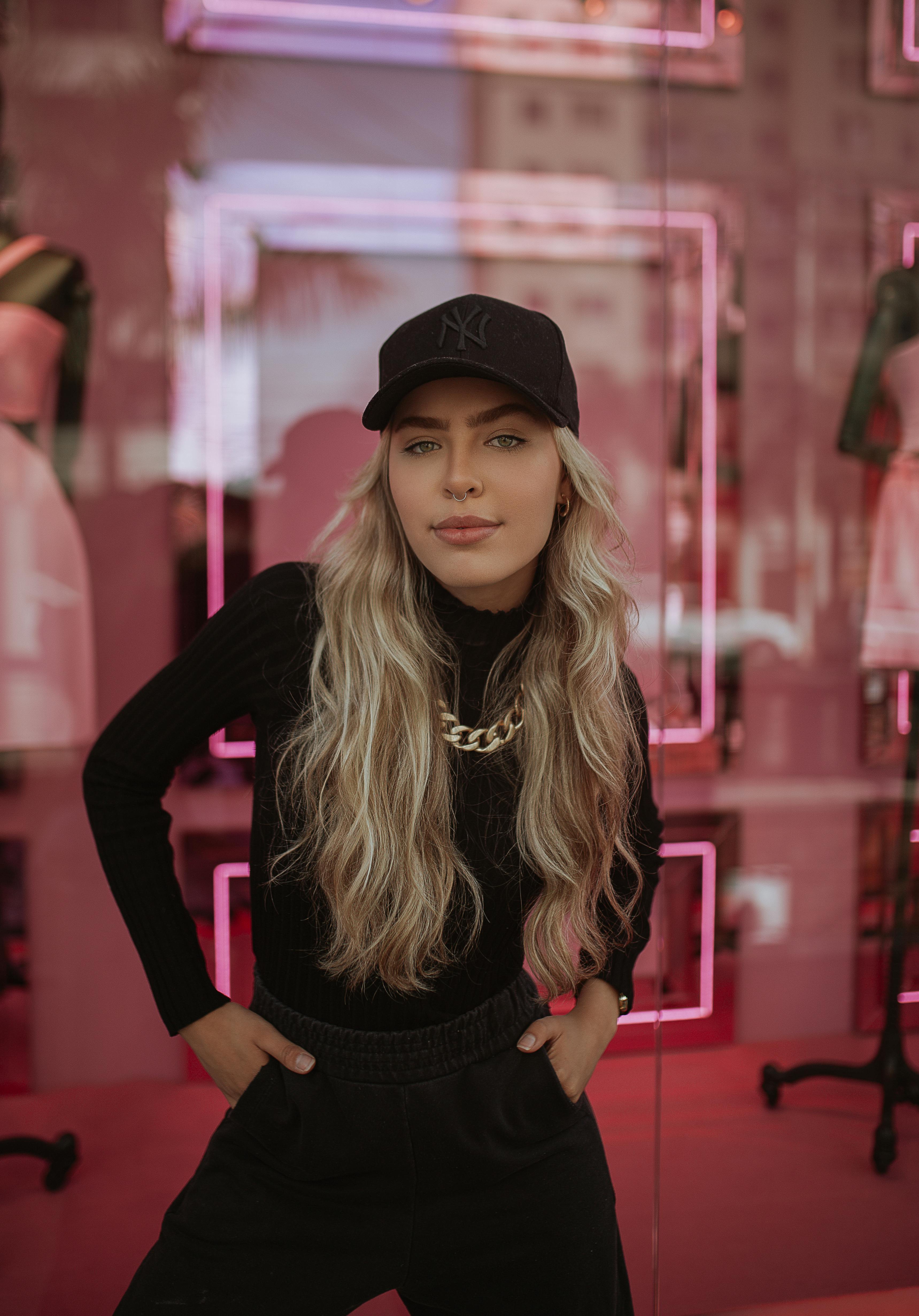 Blonde woman in fashionable attire with a baseball cap poses confidently in a vibrant urban setting.