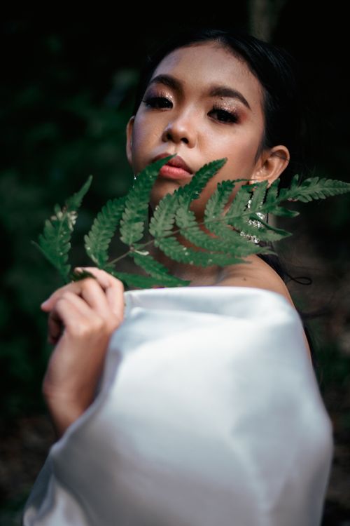 Free A young woman in a white silk garment holding fern leaves in a serene forest setting. Stock Photo
