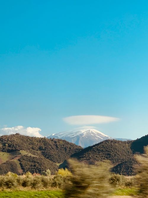 Free A scenic view of a snow-capped mountain under a bright blue sky with surrounding forested hills. Stock Photo