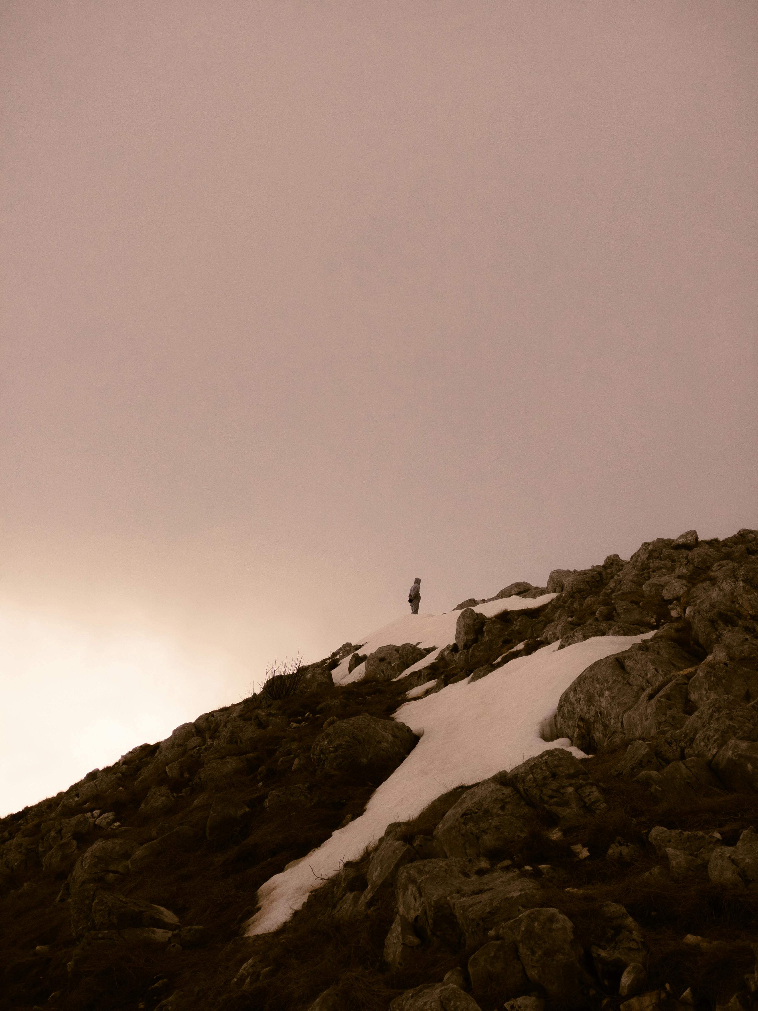 A lone hiker stands atop a rocky snow-covered mountain peak at dusk, under a serene, overcast sky.