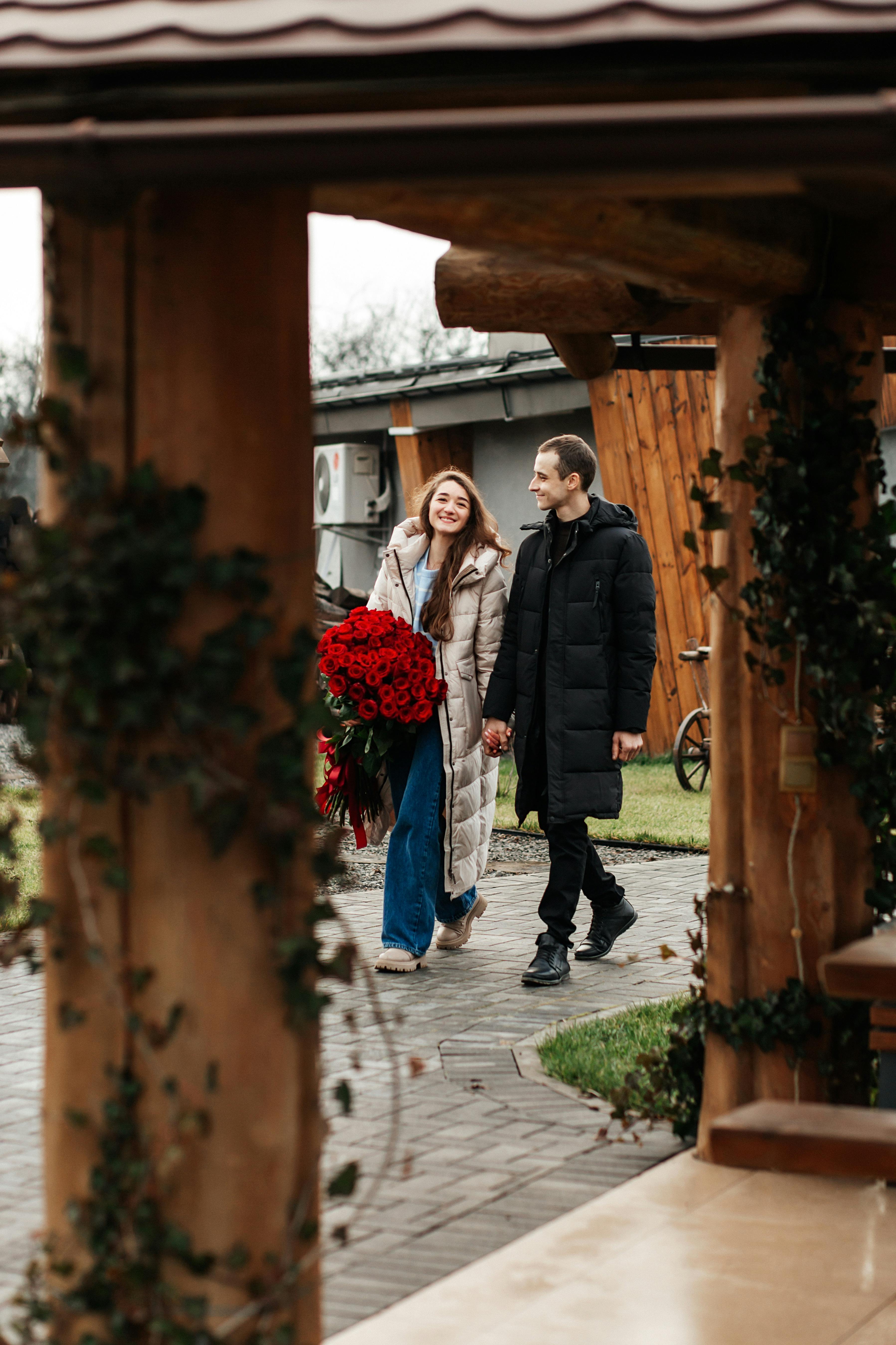 A happy couple walks outdoors holding hands with a bouquet of red roses on a chilly day.