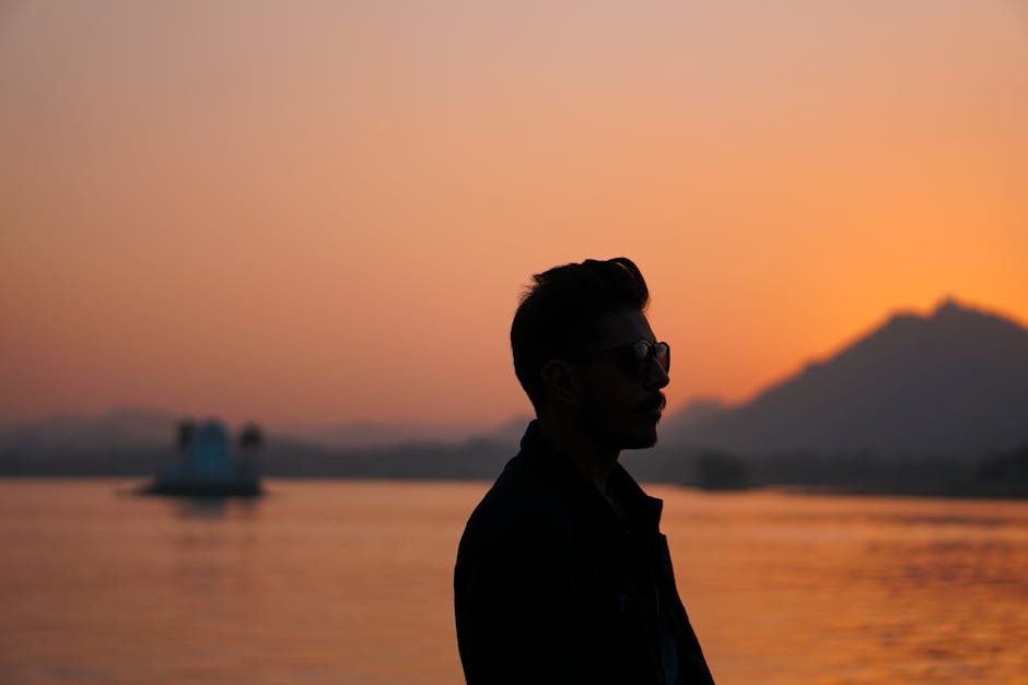 Silhouette of a man wearing sunglasses during a serene sunset by the sea, with mountains in the background.