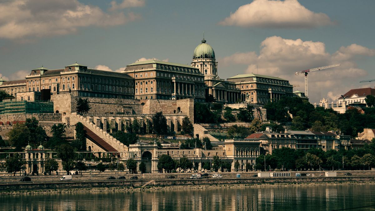 Scenic view of Buda Castle in Budapest with the Danube River in the foreground under a partly cloudy sky.