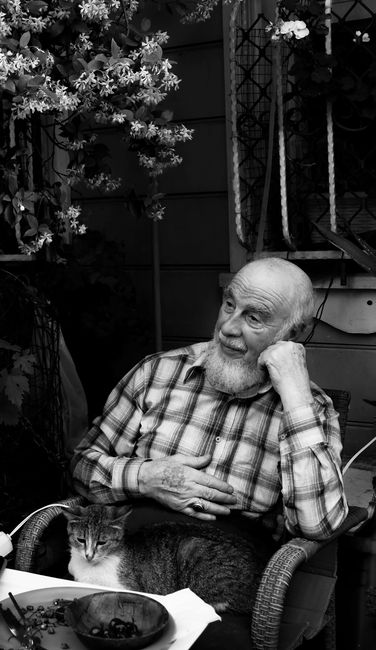 A senior man sitting with a cat on his lap in a garden, black and white.
