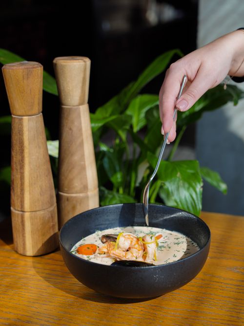 Free A hand holding a spoon over a bowl of creamy seafood soup with shrimps, herbs, and vegetables. Stock Photo