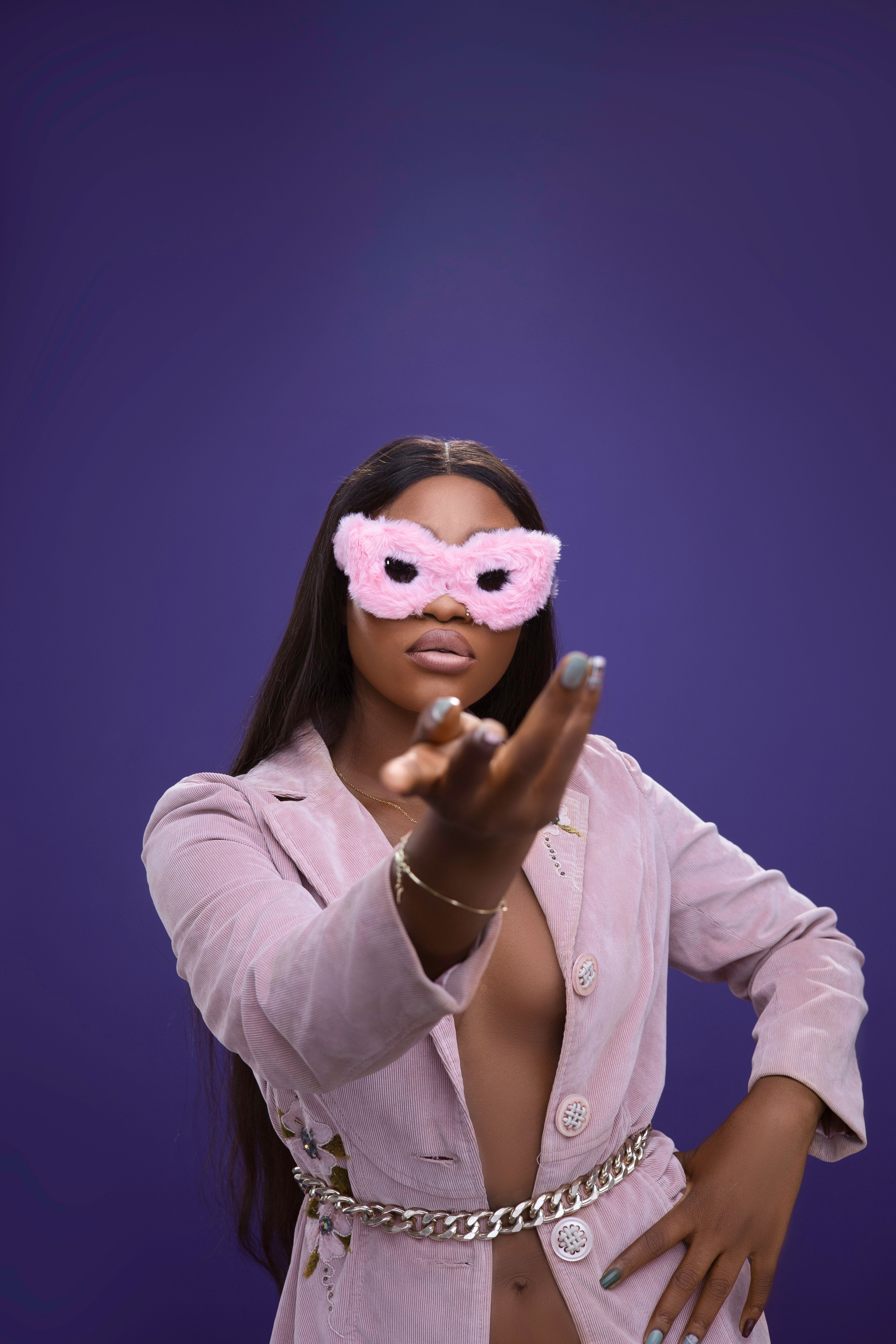 A stylish woman wearing a pink mask and jacket poses confidently against a purple backdrop.