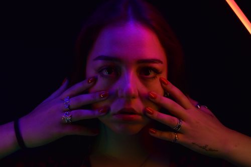 Free Close-up portrait of woman in dramatic multicolored lighting, framed by hands adorned with rings. Stock Photo
