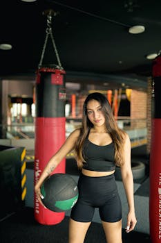 Athletic woman in gym wearing sportswear, holding a medicine ball, ready for workout with punching bags around.
