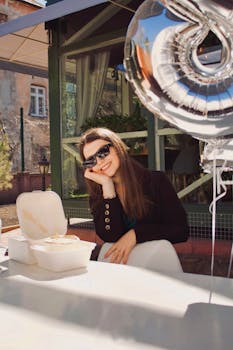 Smiling woman in sunglasses celebrates outdoors with cake and silver balloons.