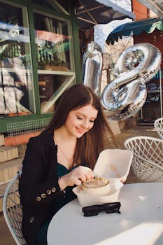 A young woman celebrating her birthday with a cake at a sidewalk cafe, smiling with joy.