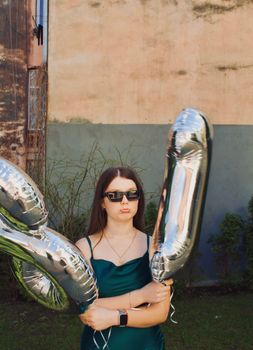 Young woman in sunglasses poses with balloons celebrating her 18th birthday outdoors.