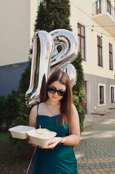 Stylish young woman celebrating her 18th birthday outdoors with balloons and cake.