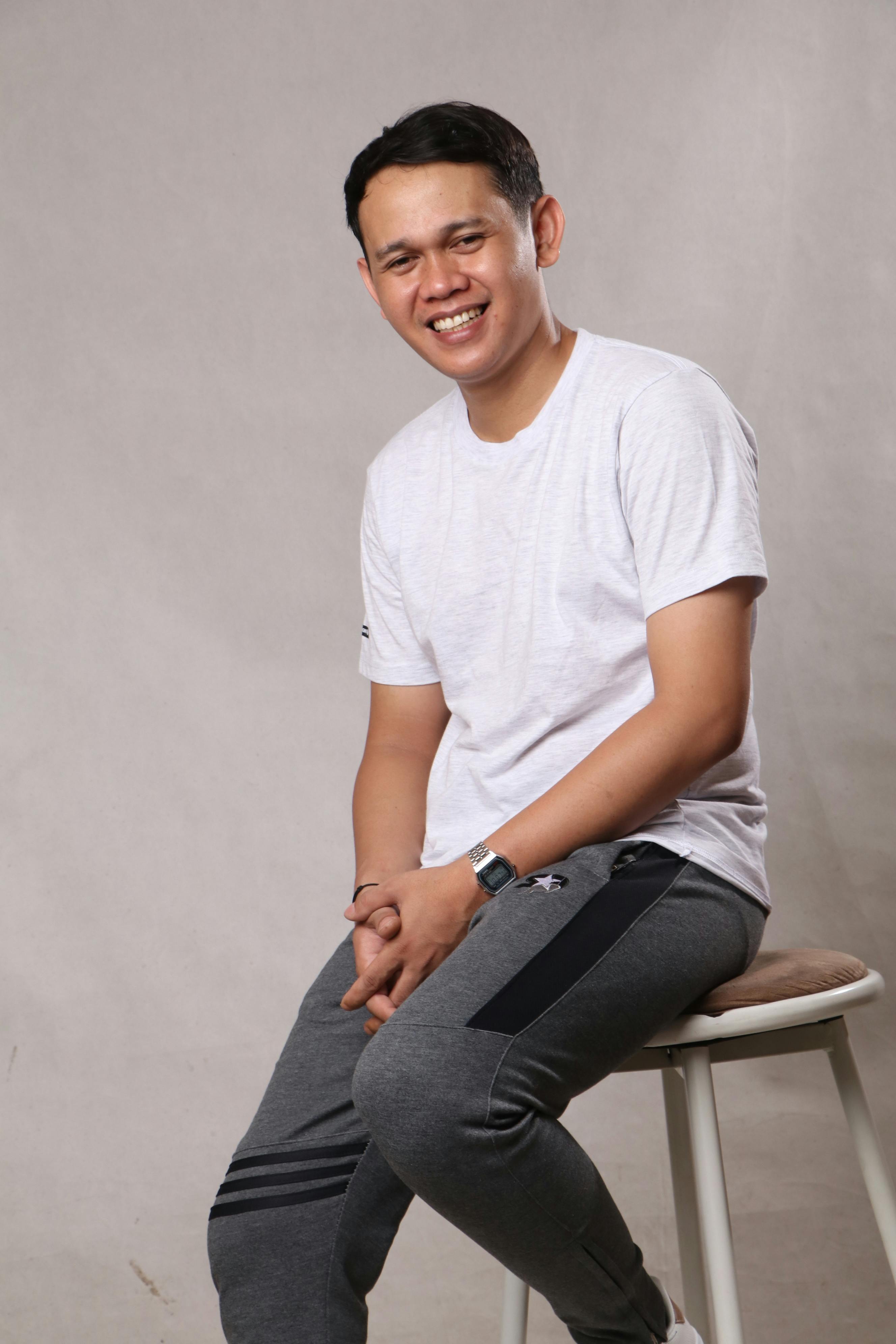Smiling young man in casual outfit sitting on a stool in a studio setting.