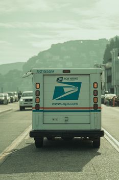 A USPS delivery truck driving down a scenic road in Sausalito, California, featuring hillside views.