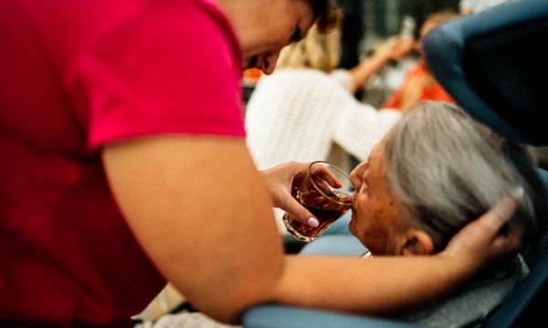 Elderly Care Worker — A caregiver helps an elderly woman drink tea, showcasing empathy and support.
