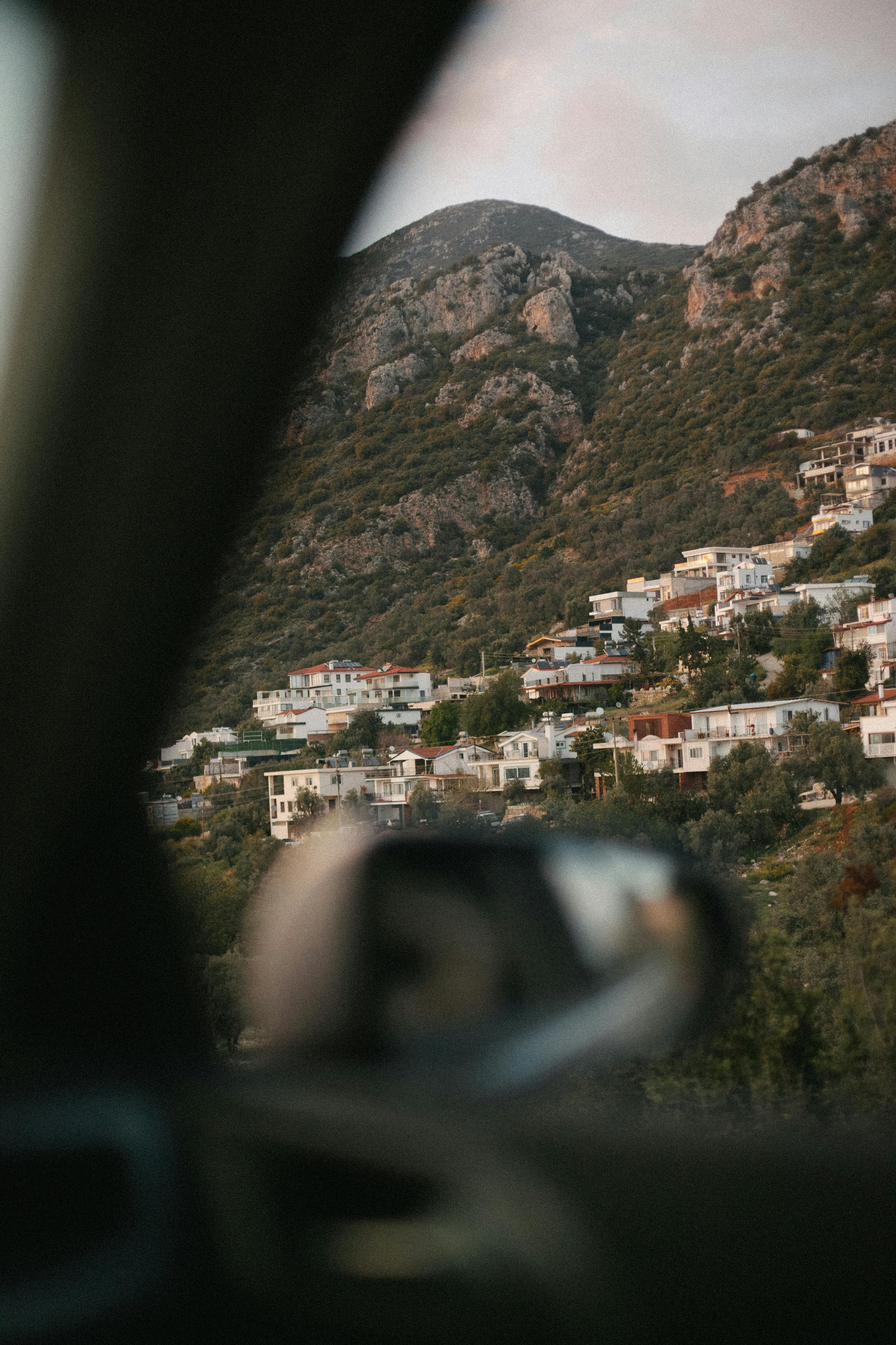 A serene hillside village seen through a car window, nestled against a rugged mountain.