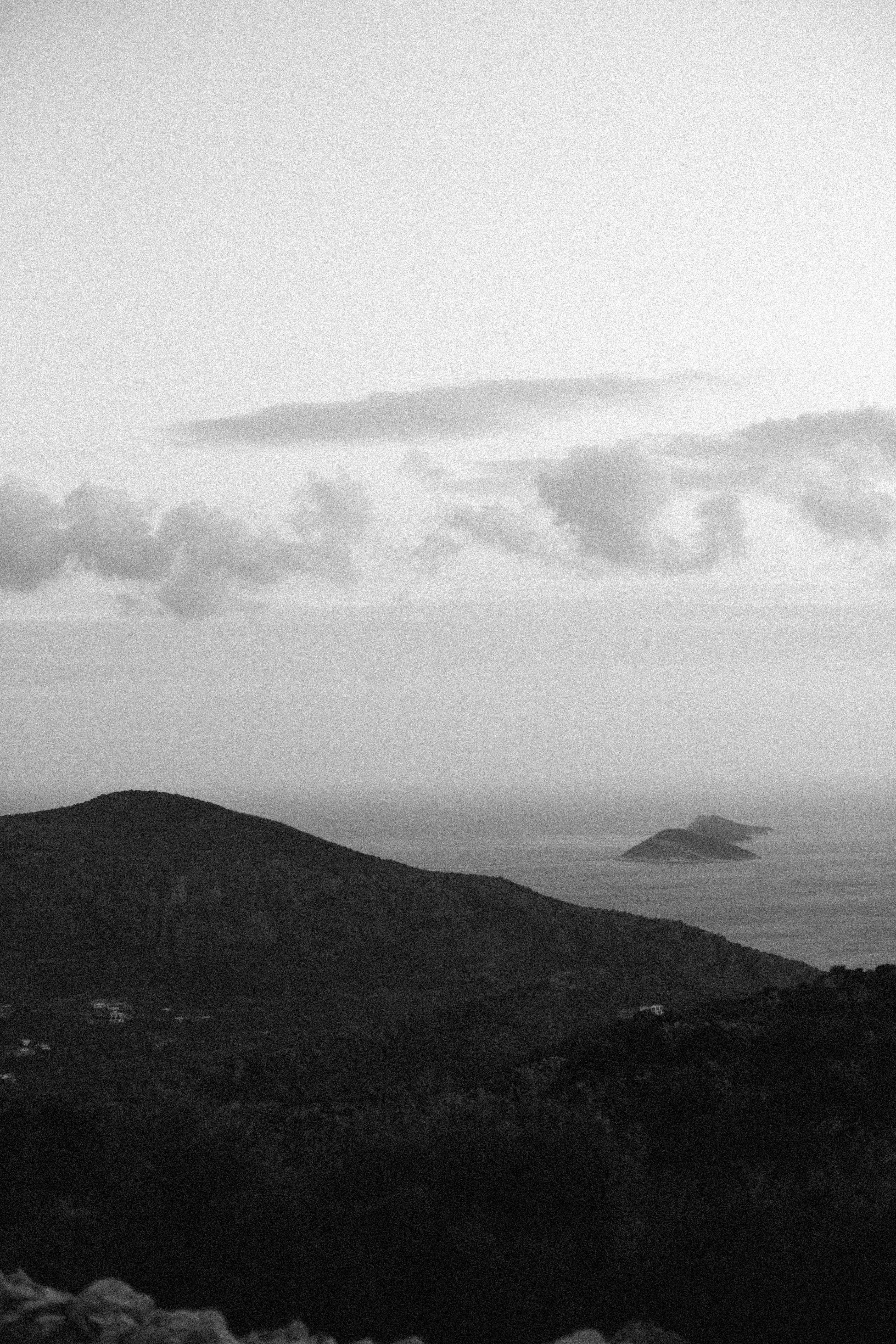 A serene black and white seascape with hills, sea, and distant islands under a cloudy sky.