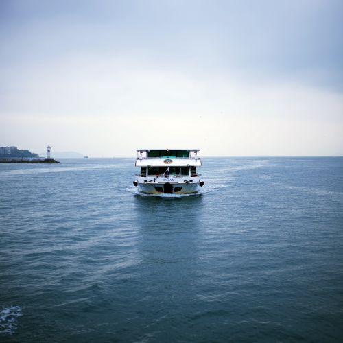 Free A ferry boat navigates the calm sea with a lighthouse in the distance, under a cloudy sky. Stock Photo