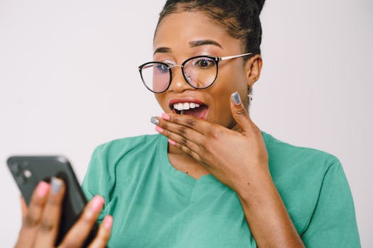 A young woman in glasses looks surprised while using her smartphone indoors.