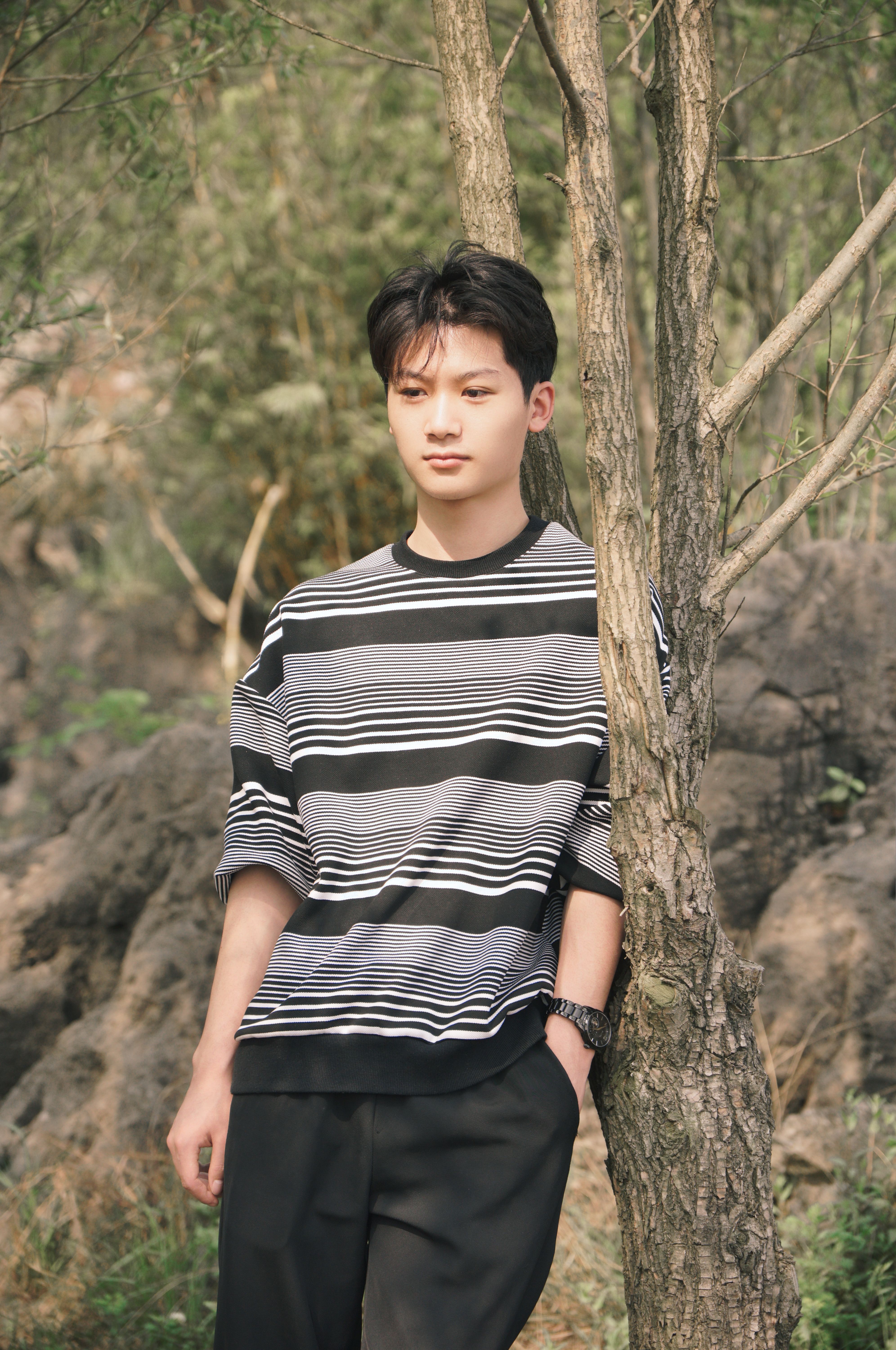 Teenage boy in a striped sweater posing outdoors against a tree, casual fashion shot.