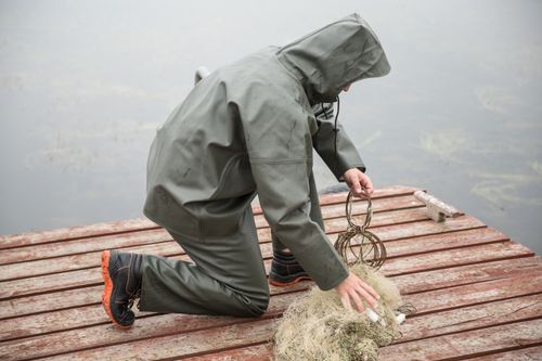 Free A person in a green raincoat kneeling on a wooden jetty picking up a fishing net. Stock Photo