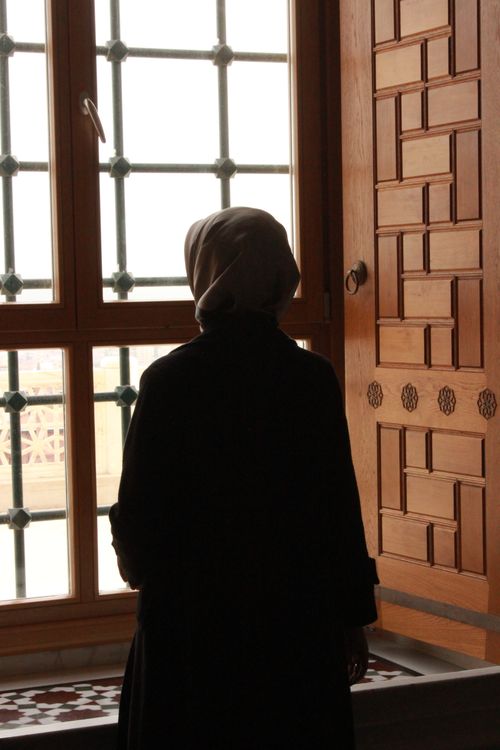 Free A woman in a headscarf stands in silhouette, gazing out a window with ornate bars. Stock Photo