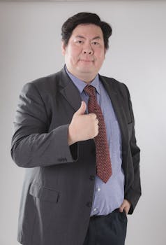 Mature Asian businessman in a suit with a thumbs-up gesture indoors.