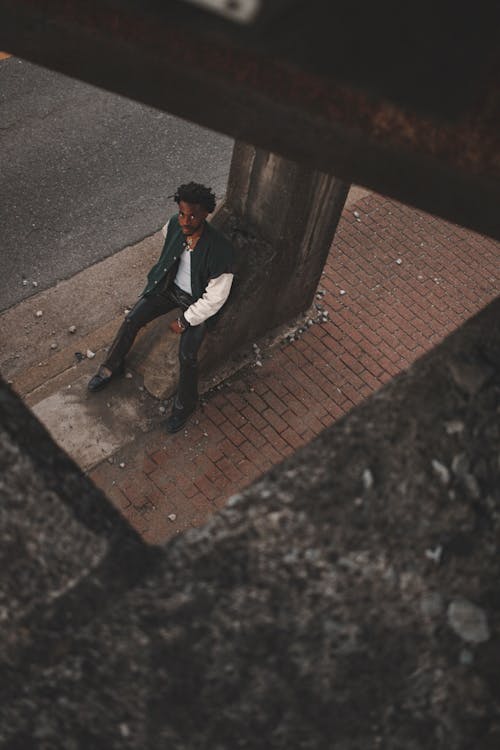 Young Man Sitting on a Concrete Pole on a Sidewalk in City 