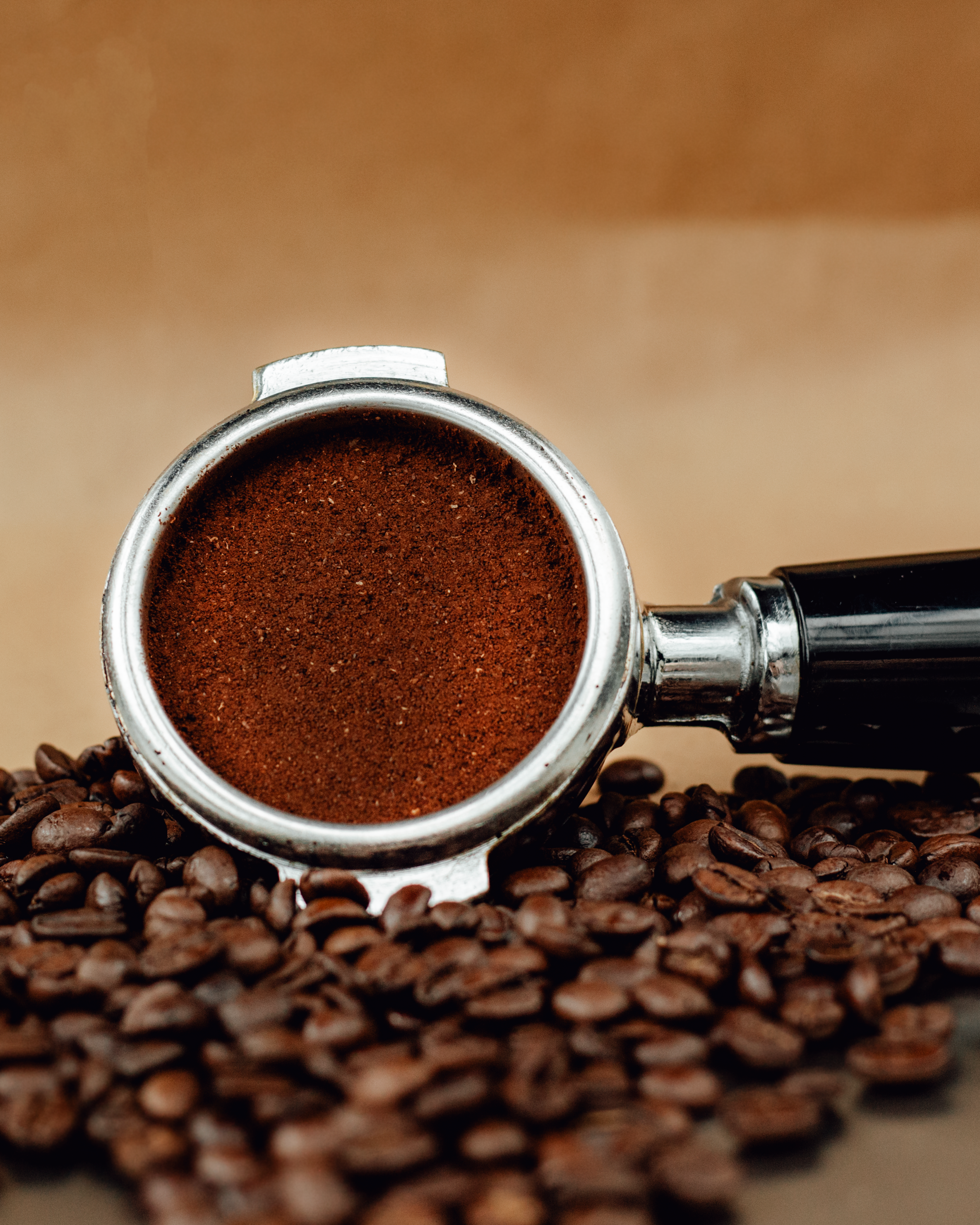Detailed shot of a coffee portafilter filled with grounds and scattered coffee beans on a brown background.