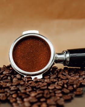 Detailed shot of a coffee portafilter filled with grounds and scattered coffee beans on a brown background.