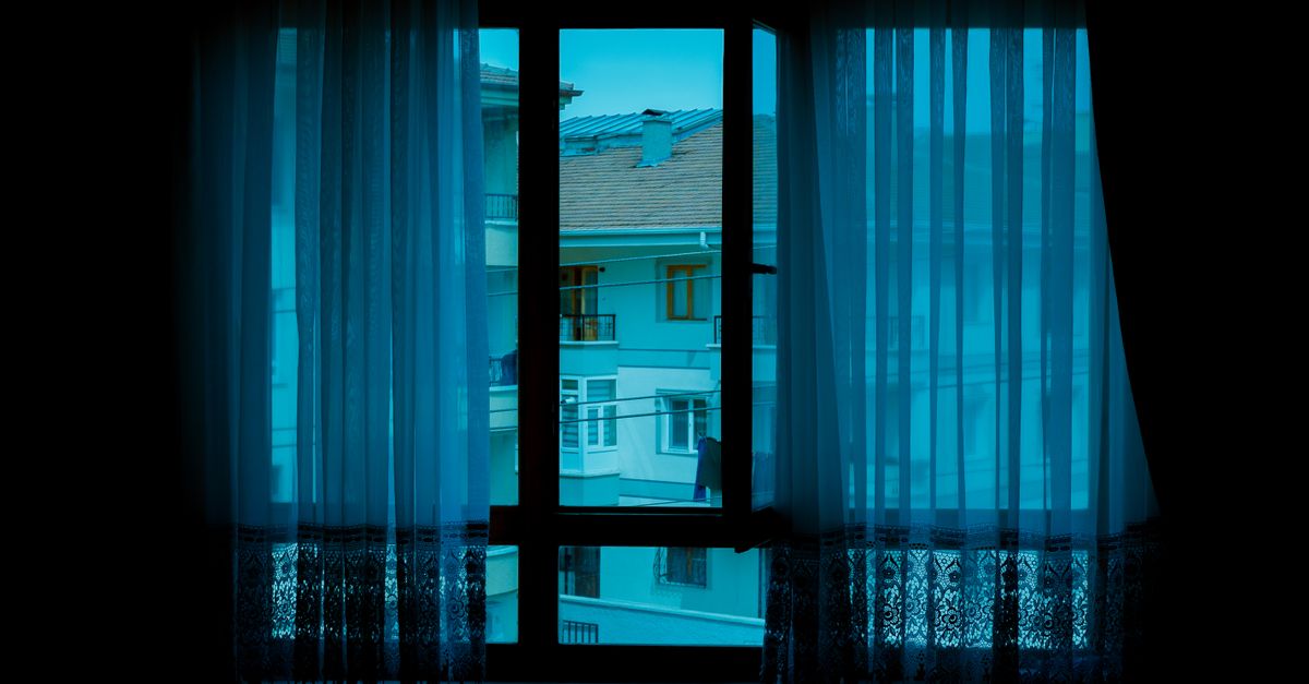 Vista de edifícios de apartamentos adjacentes através de uma janela com cortinas leves, criando uma cena urbana tranquila.
