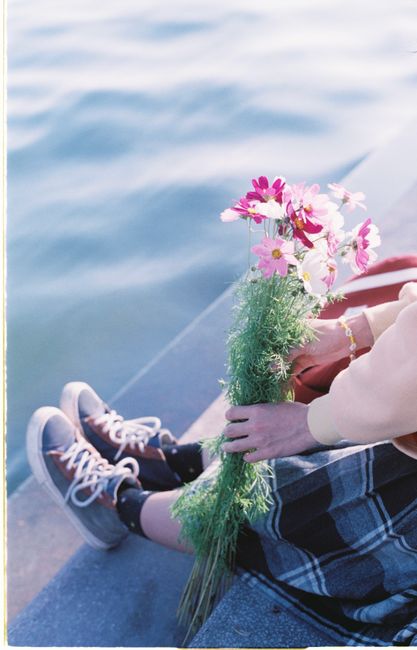A woman sits by the water holding a bouquet of wildflowers, embracing a serene moment.