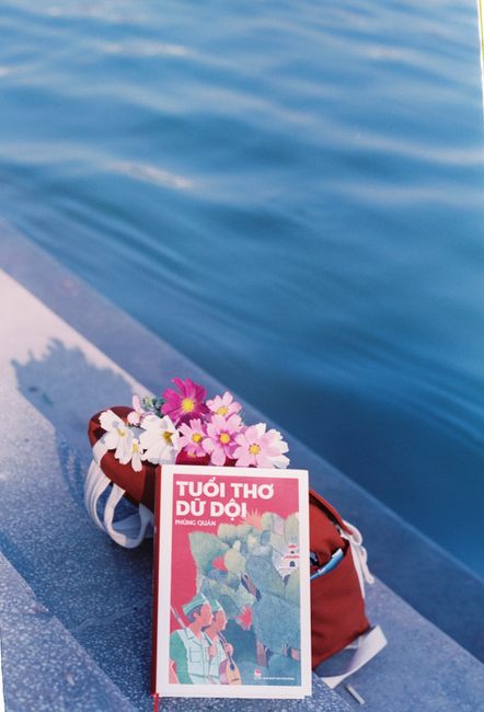 A vibrant book and flowers rest in a backpack by the serene waterside.
