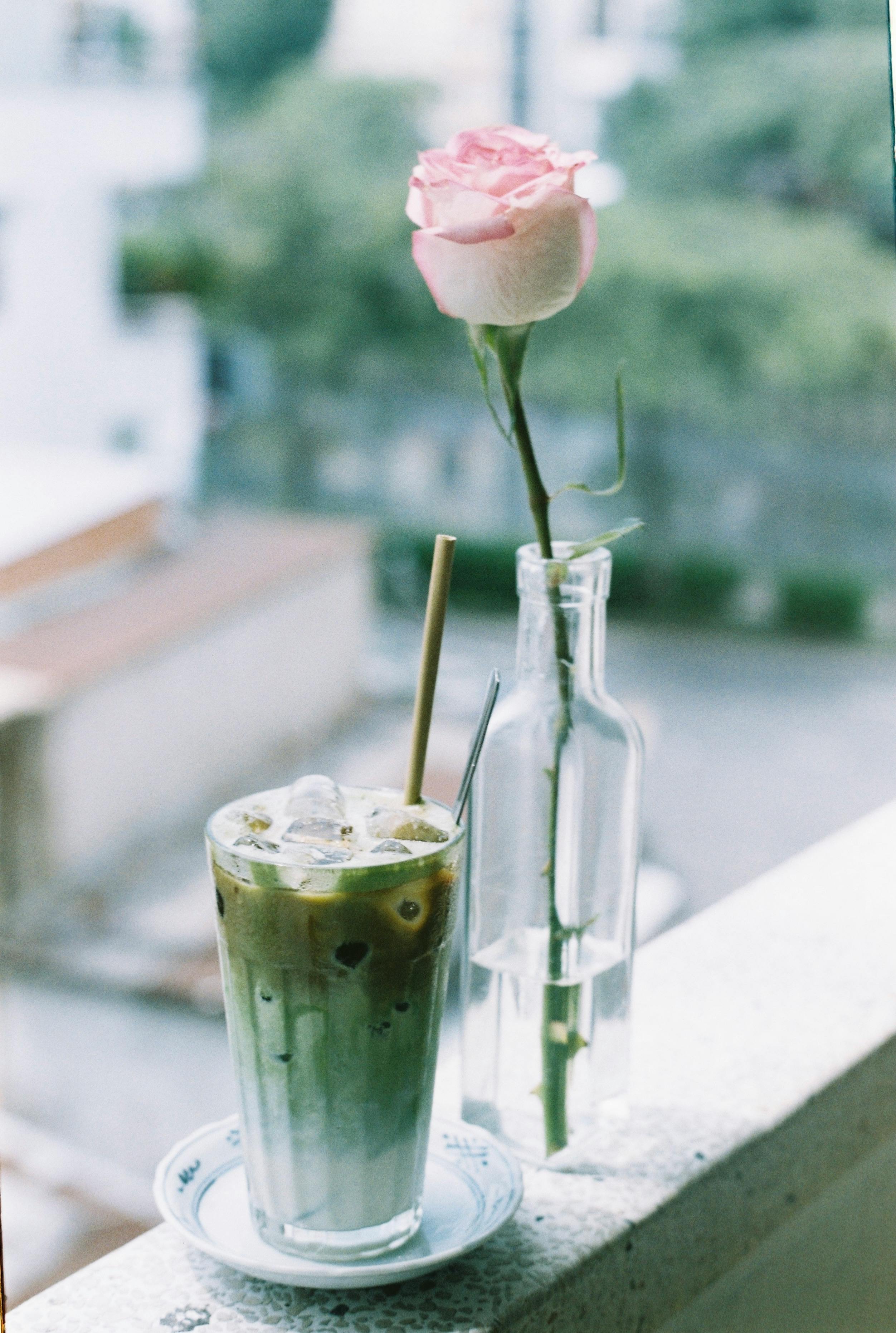 Aesthetic shot of iced matcha latte and pink rose in a vase on a ledge.