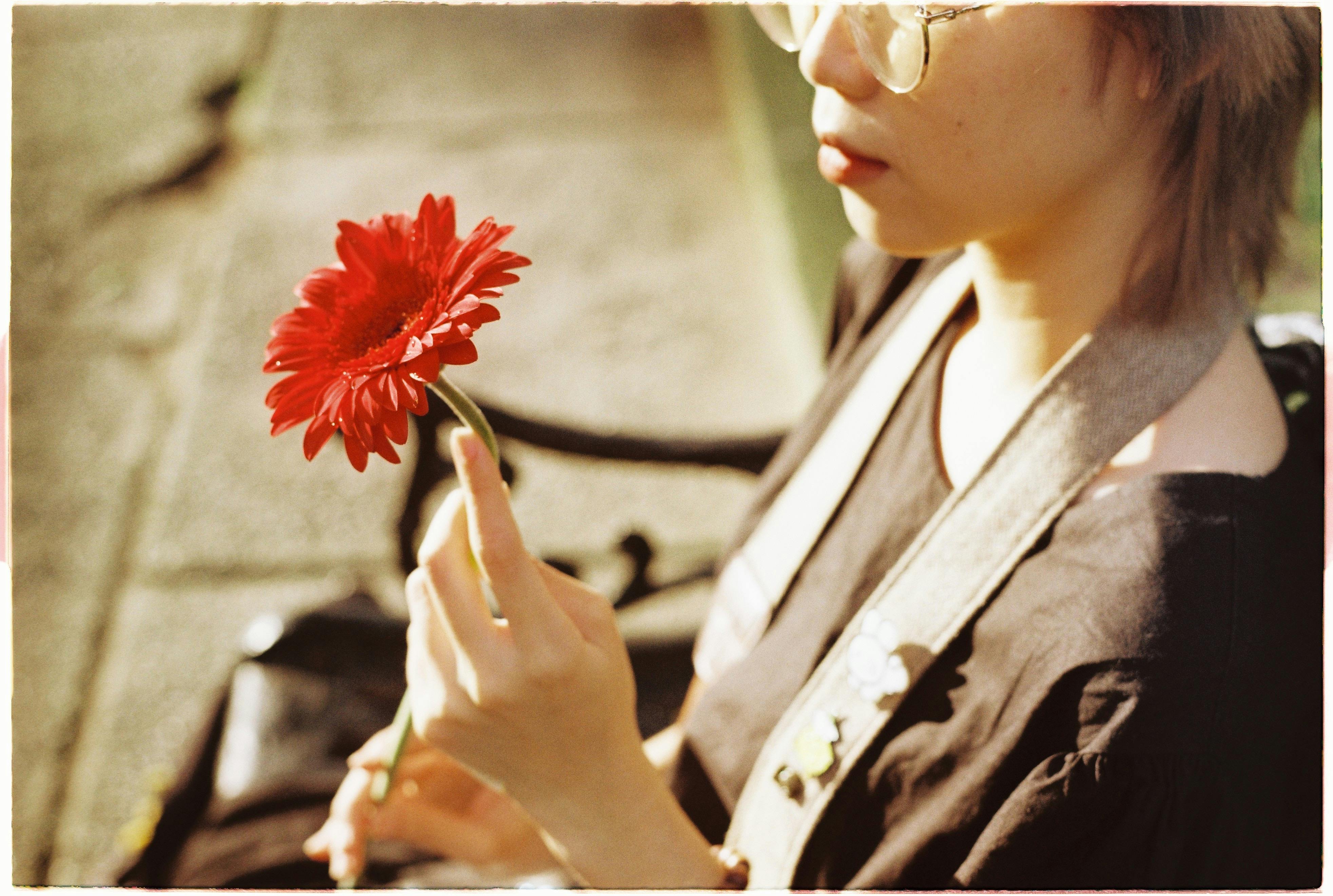 A woman sitting outdoors, holding a vibrant red gerbera daisy in soft sunlight.
