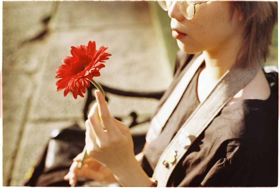 A woman sitting outdoors, holding a vibrant red gerbera daisy in soft sunlight.