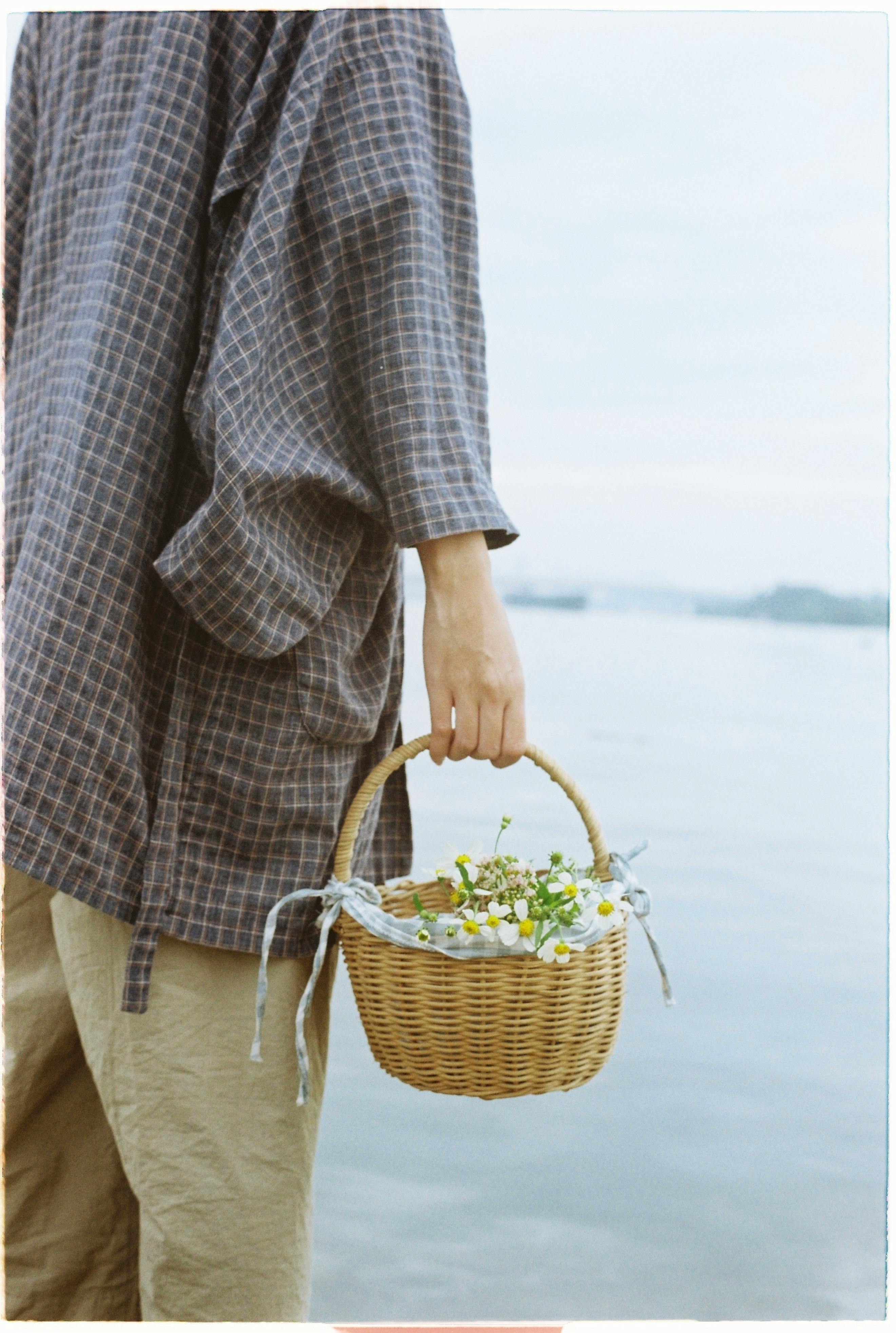 A person in casual clothing holds a wicker basket with white flowers near a body of water.