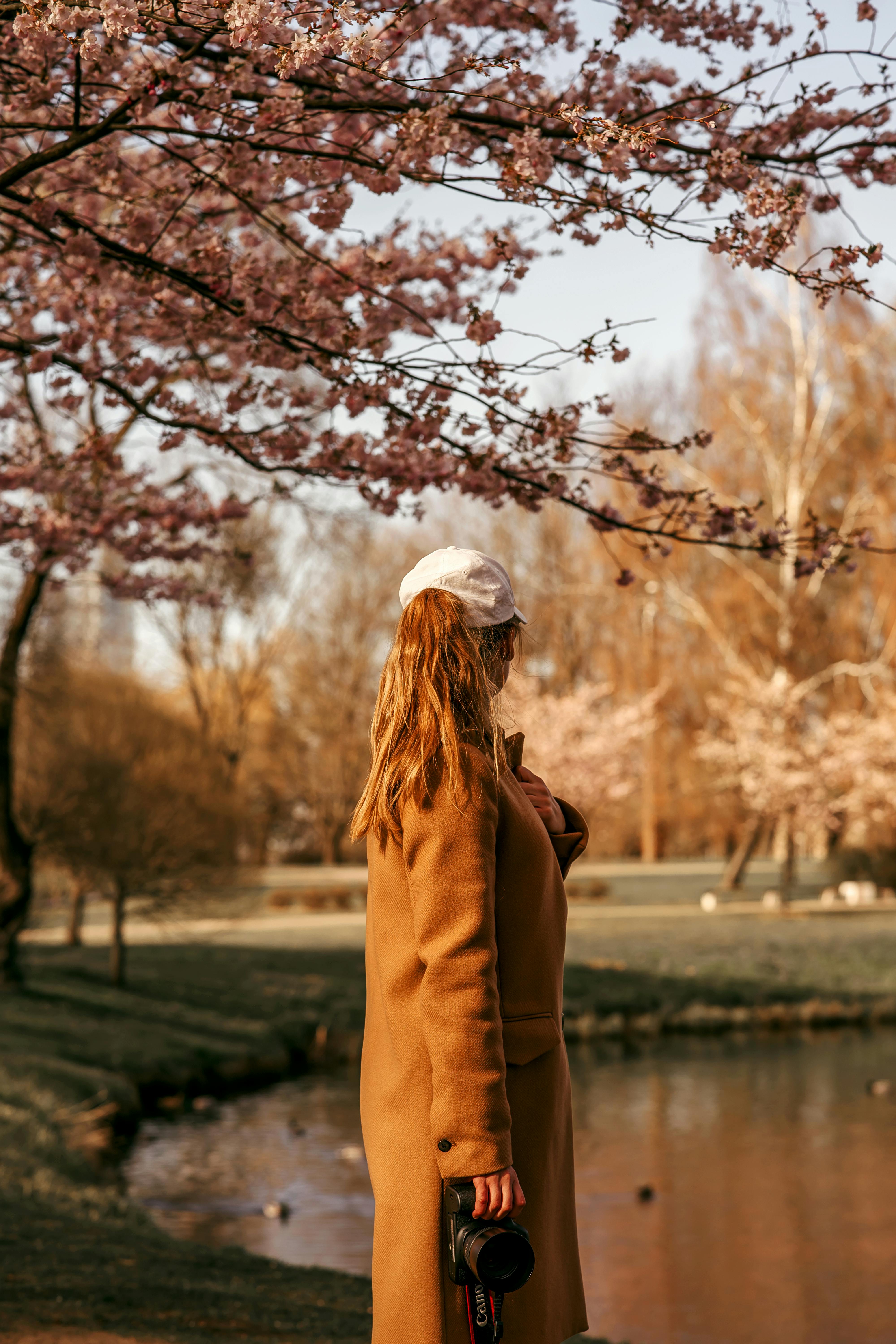 A woman in a coat holds a camera under cherry blossoms near a serene pond in autumn.