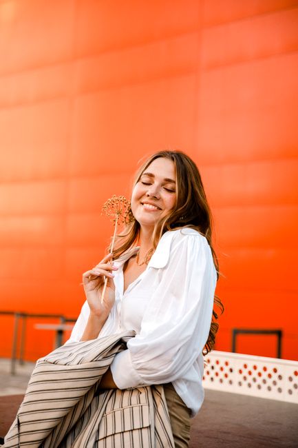 Woman smiling with eyes closed, posing against a vibrant orange background holding a flower.