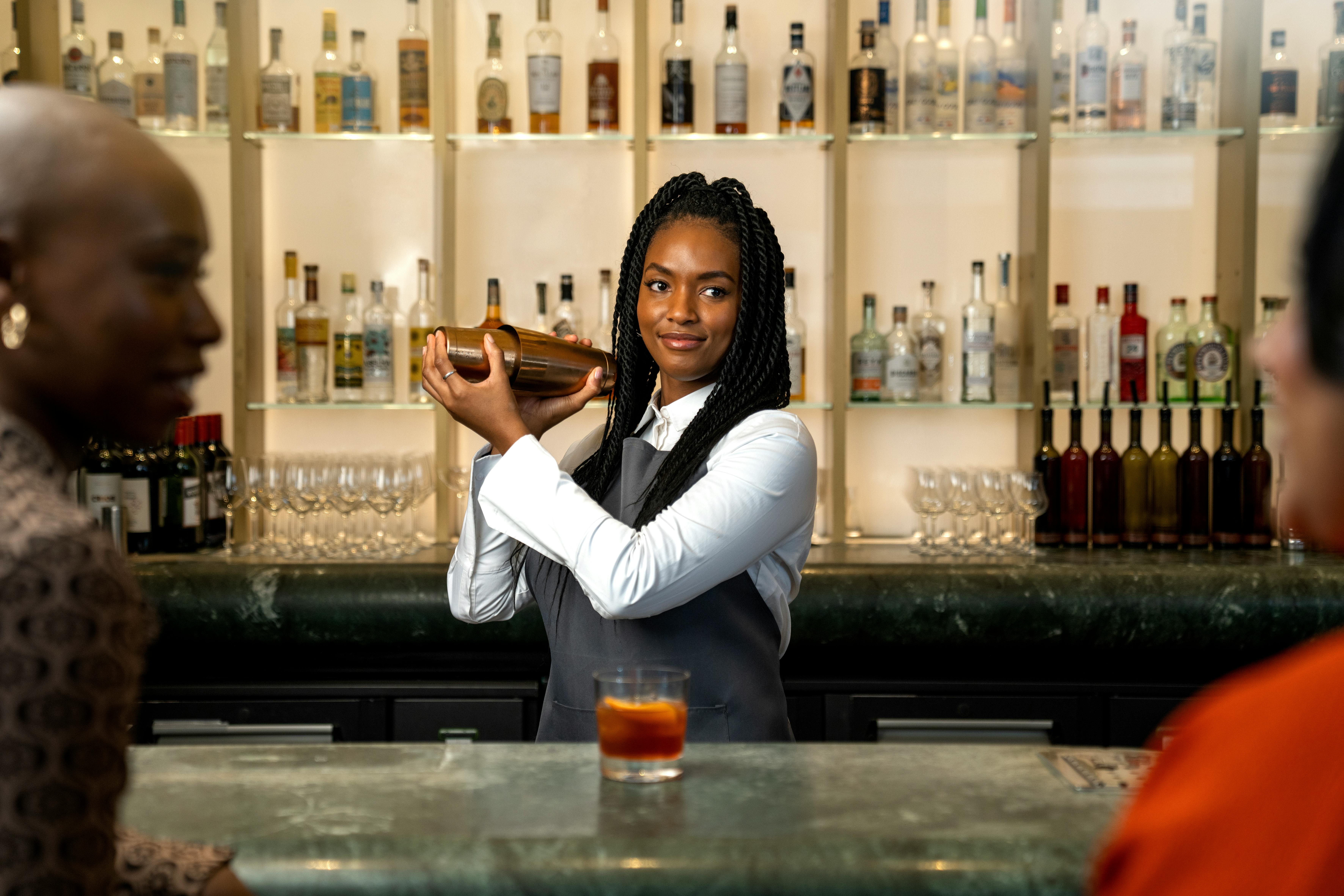 A friendly bartender with long hair mixes a cocktail for patrons at a stylish bar.
