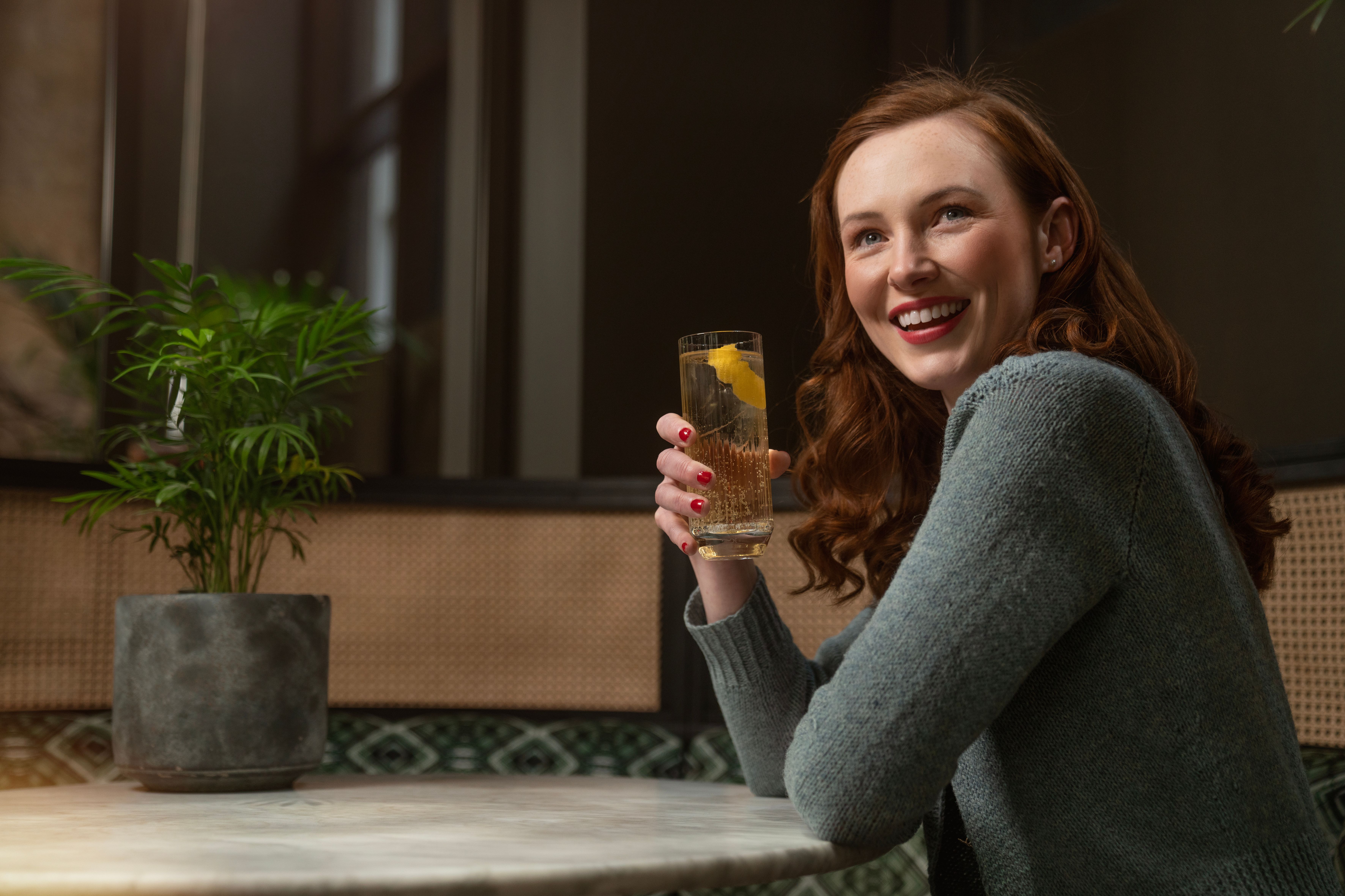 Portrait of a smiling woman with red hair holding a drink in a cozy indoor setting with a plant background.